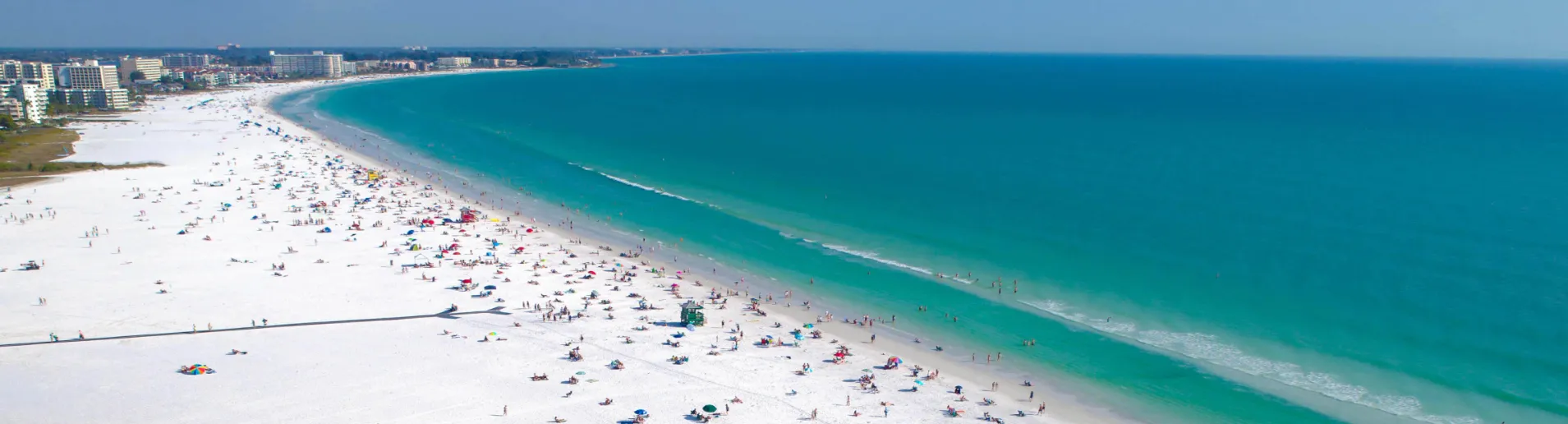 Aerial view of a beach with a lot of people sitting on the sand and swimming in the ocean. There are also some palm trees and buildings in the background