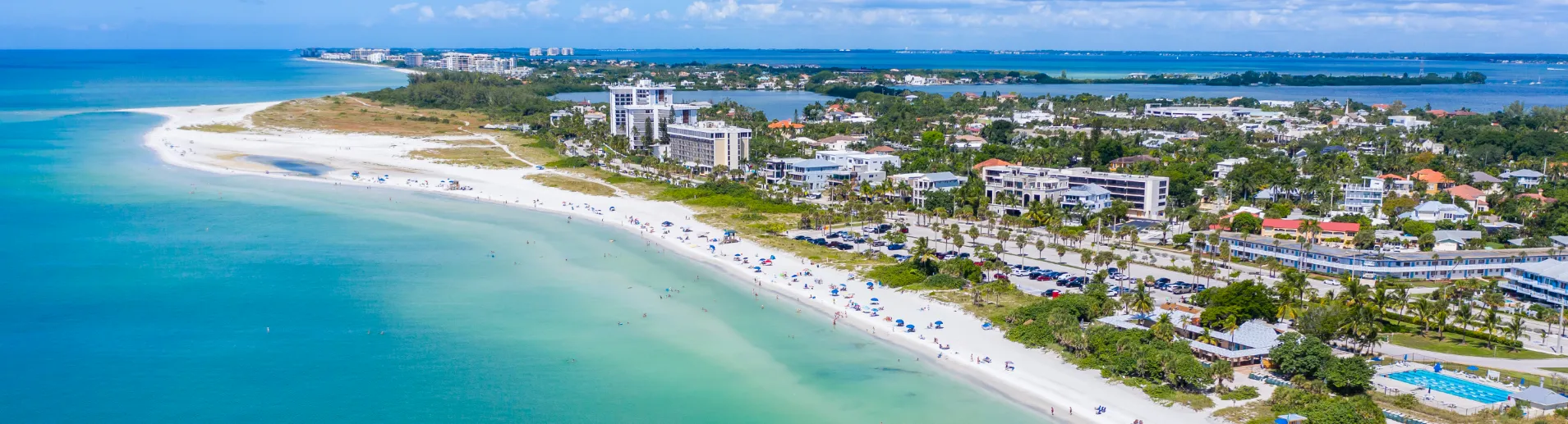 Aerial view of a beach with a lot of people sitting on the sand and swimming in the ocean. There are also some palm trees and buildings in the background